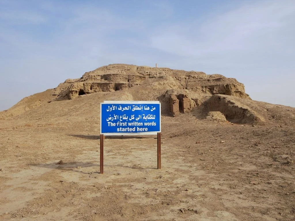 A large platform of mud bricks, eroded and slumping through time, with sloping sides and what look like they were staircases ascending its sides. A blue sign with white lettering in Arabic and English is planted in the foreground. It reads: The first written words started here