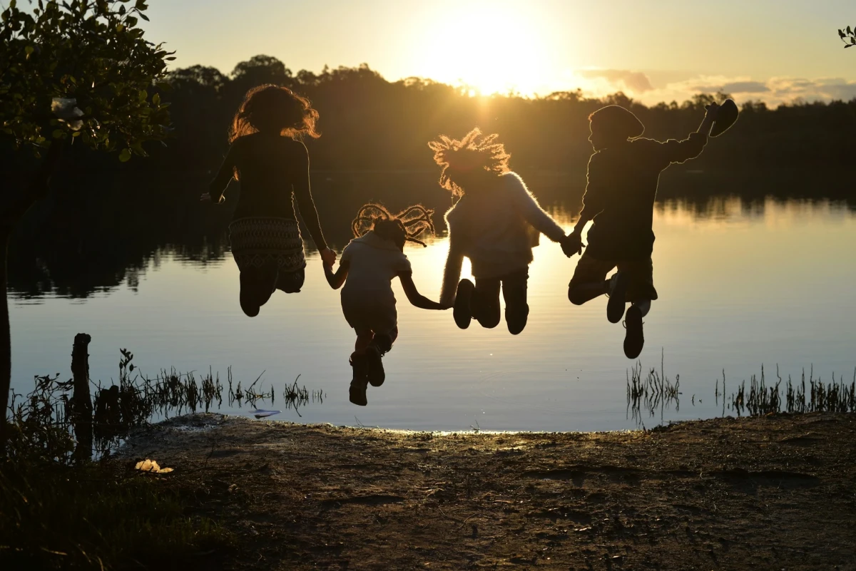 Four children, holding hands, are jumping high into the air at the edge of a small lake, their hair flying. The water in front of them shines golden in the setting sun. They are silhouetted against the sunset.