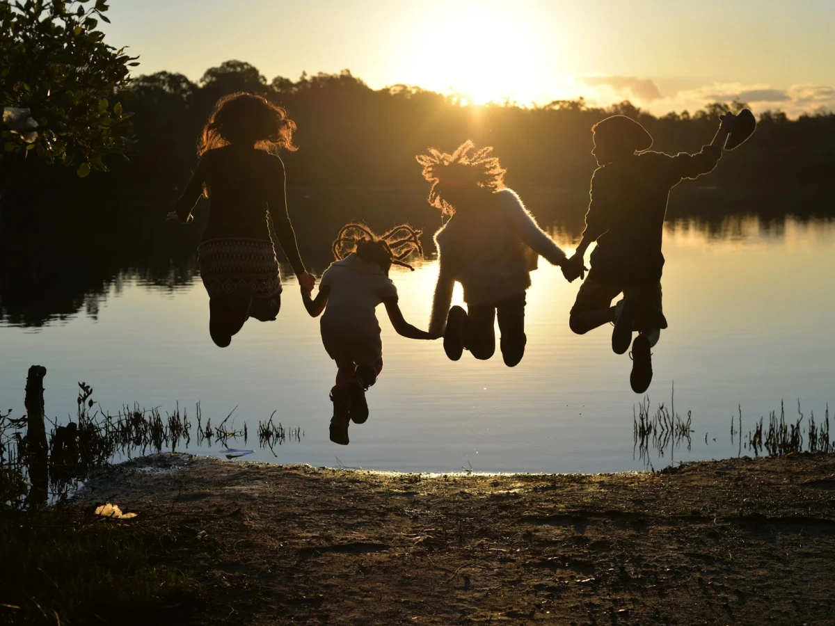 Four children, holding hands, are jumping high into the air at the edge of a small lake, their hair flying. The water in front of them shines golden in the setting sun. They are silhouetted against the sunset.