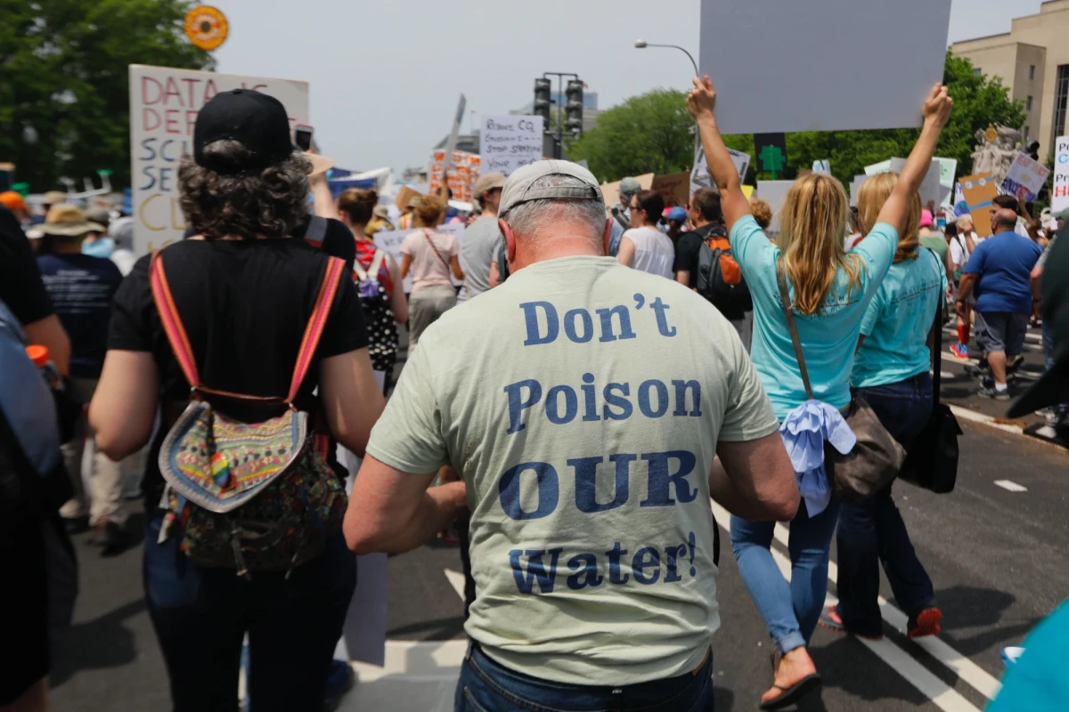 Back of a group of protesters. The man in front of the camera wears a T-shirt reading Don't Poison Our Water.