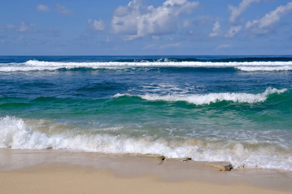 On a beach at the edge of a calm sea. In the distance a line of surf is rippling white against a deep blue ocean. In the foreground an edge of bubbling water creeps up onto the sand.