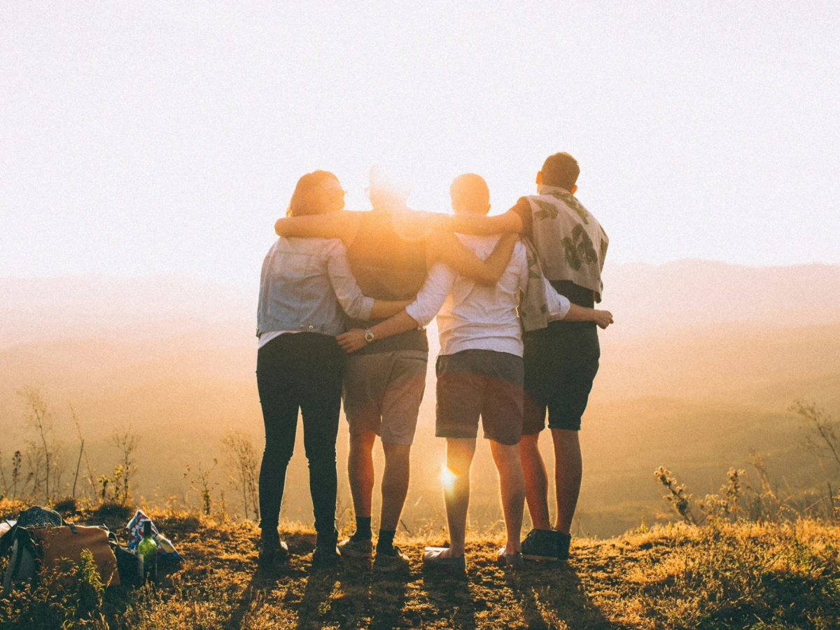 Four friends in shorts, on a picnic, are standing in a row with their backs to us, their arms slung around each other’s necks, backlit by a sun that is low in the sky. They gaze across the precipice of a hill down to a misty valley below.