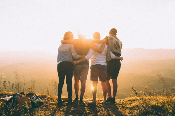 Four friends in shorts, on a picnic, are standing in a row with their backs to us, their arms slung around each other’s necks, backlit by a sun that is low in the sky. They gaze across the precipice of a hill down to a misty valley below.