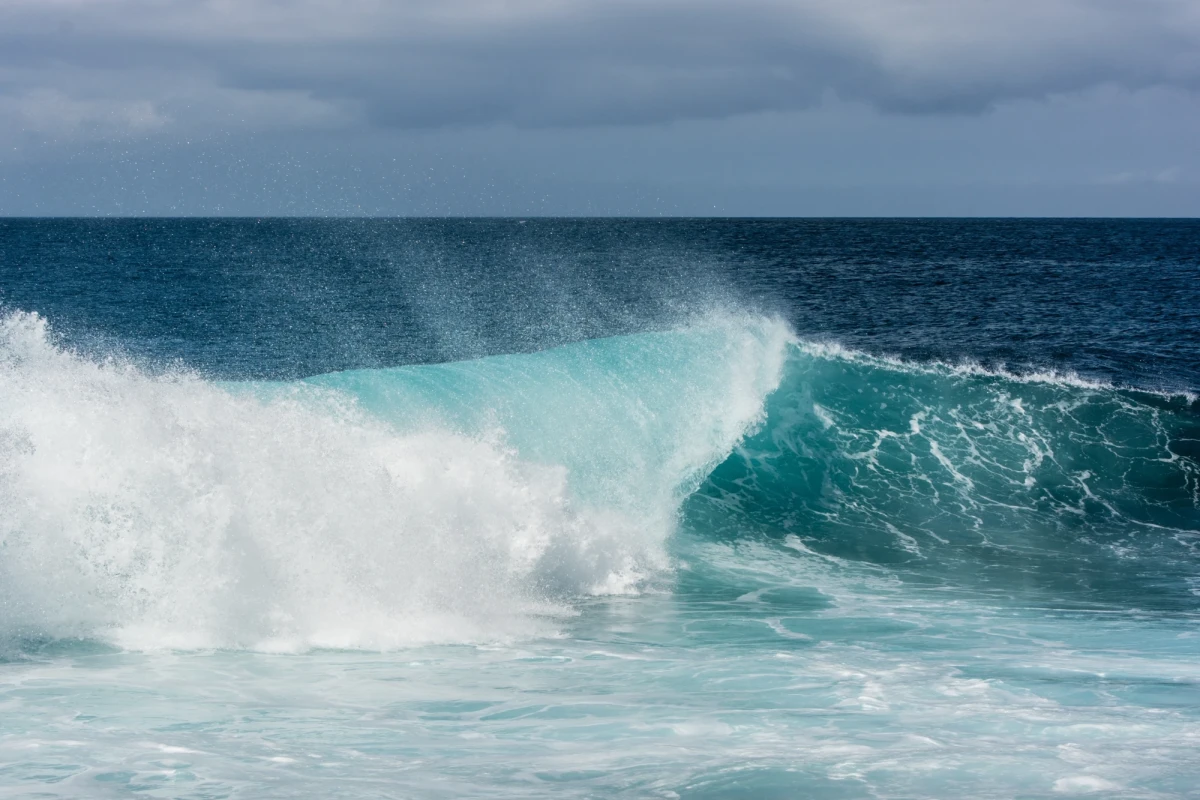 Aqua blue breaker throwing off white spray as it curls toward the surface of the sea. In the distance the ocean is deep turquoise blue under a blue sky.