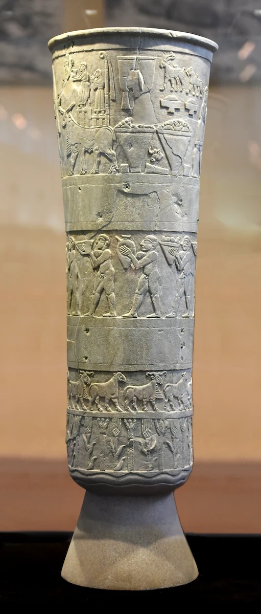 Close-up of the tall, thin Warka Vase or Uruk Vase, glowing rich amber under museum lights. Vertical sides with a slight opening toward the top, covered by carved images.