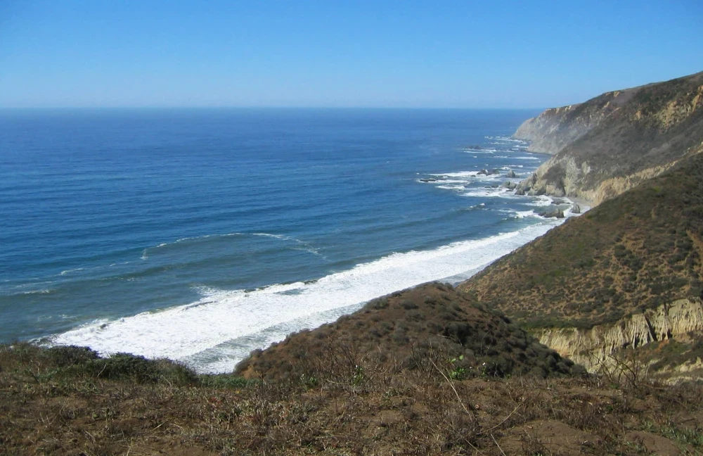 From a trail at the top of rocky seaside cliffs looking down toward the white foamy surf at the edge of the water. The deep blue rippling sea extends to forever, where it meets the lighter blue of the sky.