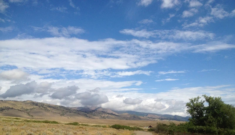 Big sky and clouds over a far horizon of brown hills. The clouds near the ridgeline are small puffs leaning right, as if moving quickly. Higher clouds are hazy and long.