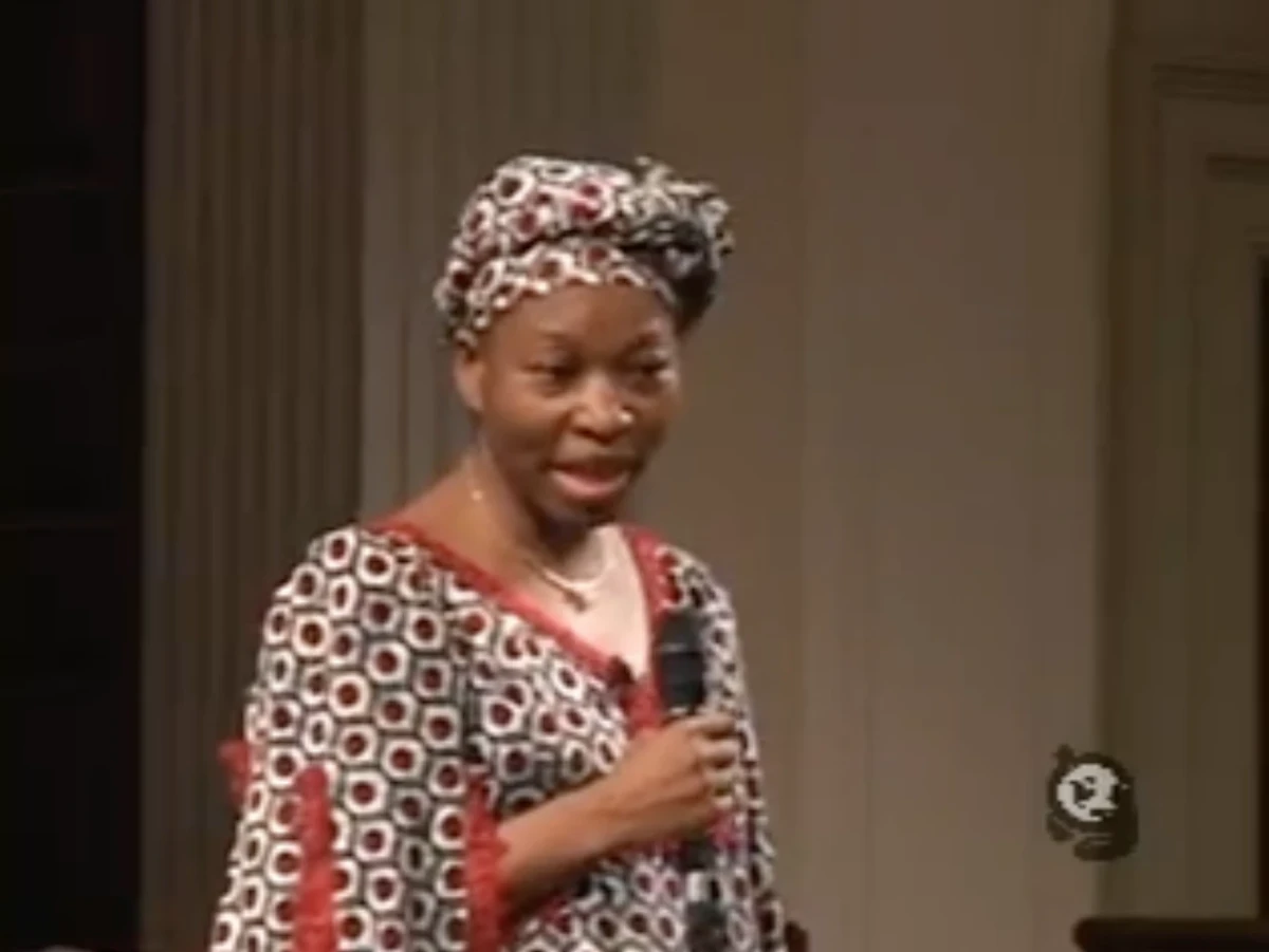 An African woman wearing a dress and matching head wrap in a fabric patterned with small hexagons stands with a microphone, talking.
