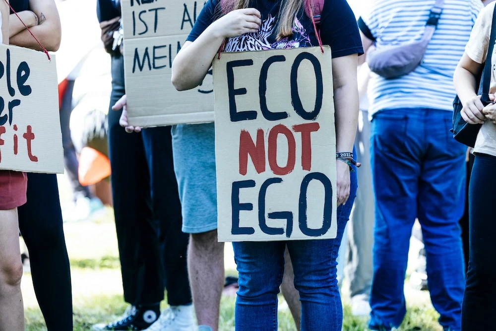 View of people standing around at a protest holding signs. The sign in front reads, ECO NOT EGO.