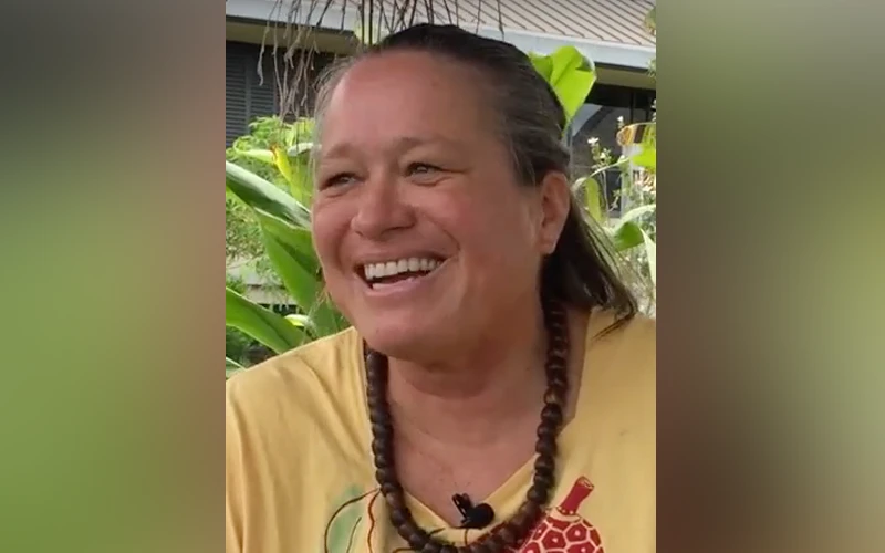 Manulani Aluli Meyer sitting outdoors with a big smile, wearing a yellow shirt and a beaded wooden necklace.