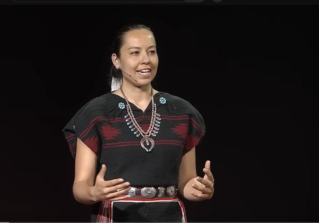 A woman stands speaking in front of a black background. She wears a black and red woven Diné dress with silver and turquoise jewelry.