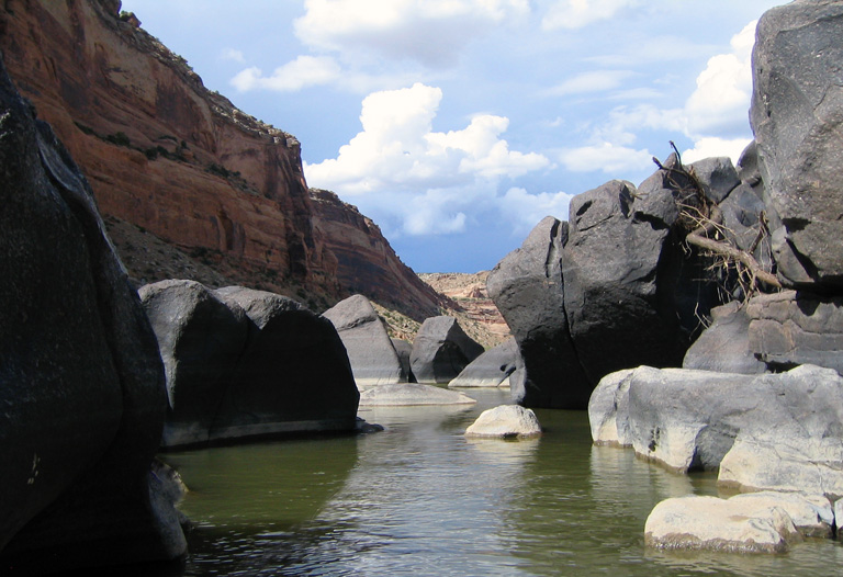 On the surface of a still dark river passing between huge gray boulders, framed by red rock canyon walls. Fluffy clouds float in blue above.