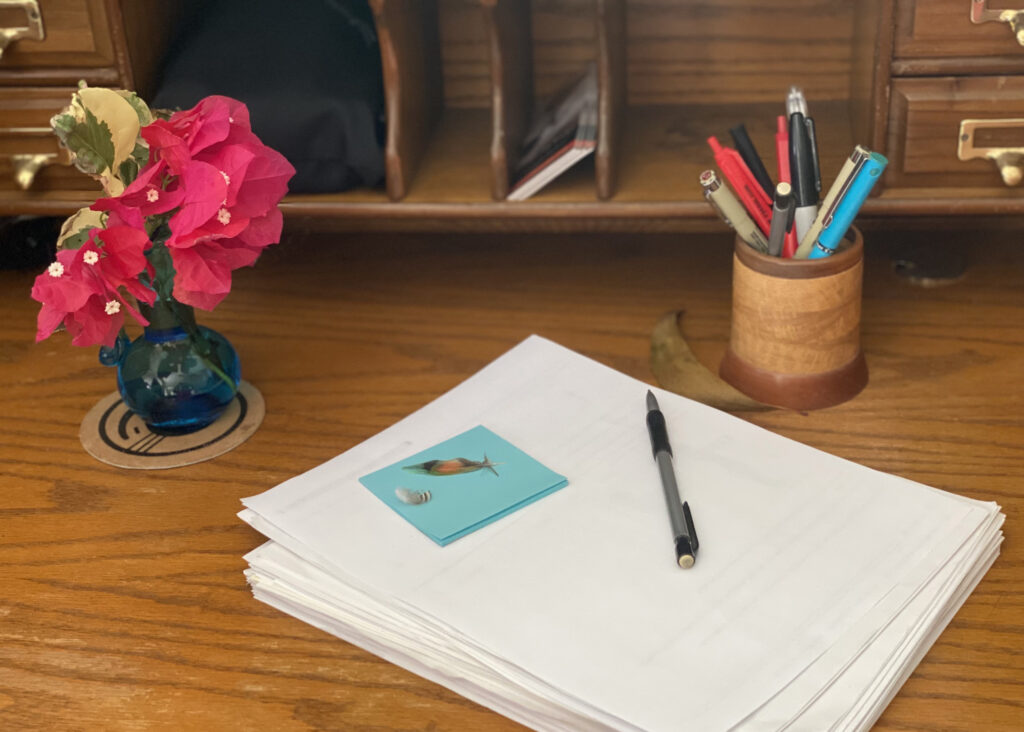 A sheaf of manuscript pages on a wooden desk with pencil, vase of flowers, and a round carved wooden pen and pencil holder. On the sheaf of papers is a nearly used-up stack of Post-It notes and two tiny feathers