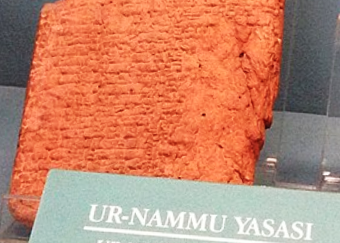 An upright slab of orange-colored clay imprinted with rows of tiny cuneiform. Slab is under glass in a museum, and the exhibit sign reads, "Ur Nammu Yasasi," or Ur Nammu Law Code.