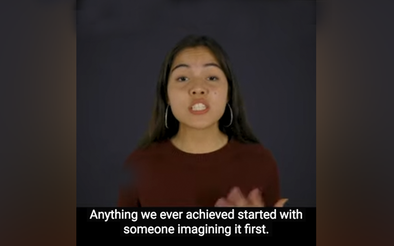 Young woman with long dark hair on a dark background sits facing the camera and saying, "Anything we ever achieved started with someone imagining it first."