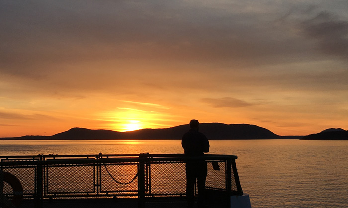 A person standing at the end of a ferry, silhouetted against a bright sunset of orange sky and orange water.