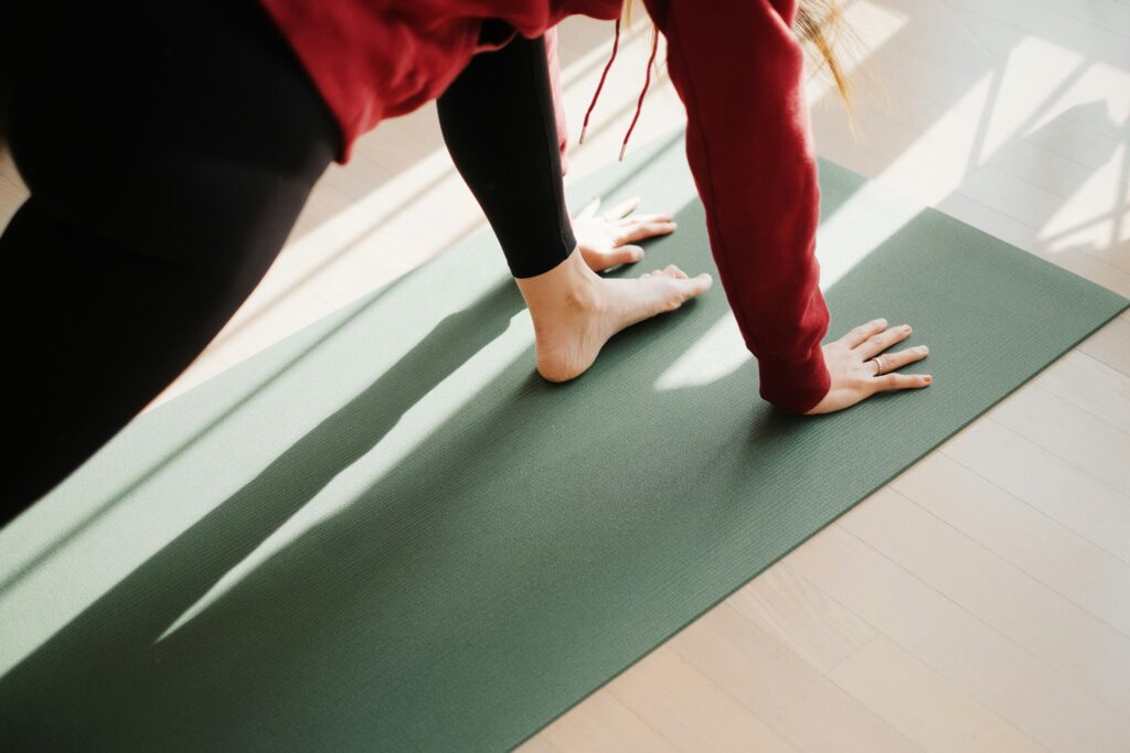 A person in a red long-sleeved sweatshirt and black leggings in a forward lunge position on a yoga mat, close-up view so that only hands and front foot and part of body are visible. Sunlight streams in across the floor and the mat.