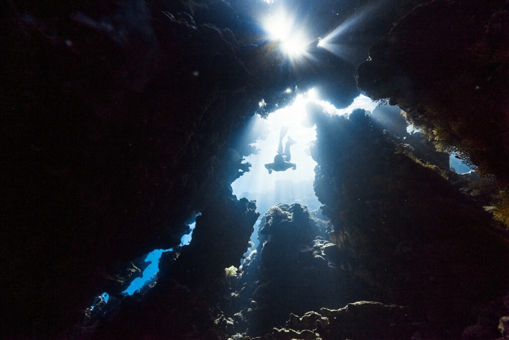 Looking up from an underwater cave at a diver swimming down into the cave. Bright sunlight streams down behind the diver.